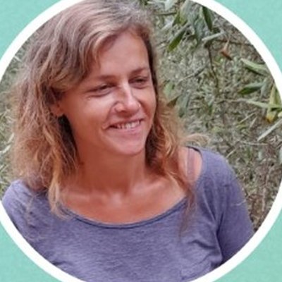 Ana Margarida Pedrosa, de cabelo ondulado aloirado e camisola lilás, sorri ao ar livre, entre ramos de oliveira.