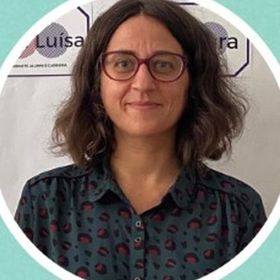 Ana Martinho, de óculos de armação escura, cabelo castanho pelos ombros e blusa estampada, sorri diante de um painel branco.