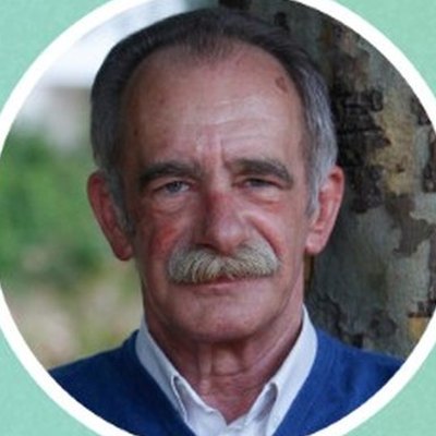 Carlos Neto, de cabelo grisalho e bigode farto, com camisola azul sobre camisa branca, sorri ao ar livre junto a uma árvore.
