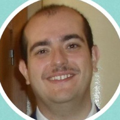 Cláudio Gomes, de cabelo curto e bigode, de fato e gravata, sorri junto a uma porta de madeira.