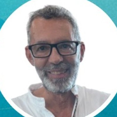 João Miranda, de óculos de armação escura, cabelo e barba grisalhos e camisa branca, sorri sobre fundo branco.
