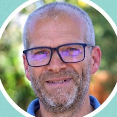 João Paiva, de óculos de armação escura, cabelo curto e barba grisalha, sorri ao ar livre, com árvores desfocadas atrás.