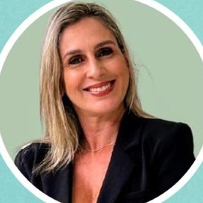 Leiriane Sanches, de cabelo louro comprido e casaco preto, sorri sobre um fundo verde-claro.