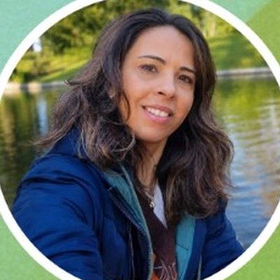Margarete Toste, de cabelo escuro ondulado e casaco azul, sorri junto a um lago num jardim.