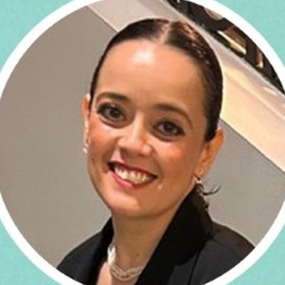 Mónica Pacheco, de cabelo escuro apanhado, colar de pérolas e casaco preto, sorri sobre fundo claro.