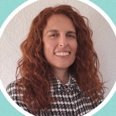 Raquel Raimundo, de cabelo ruivo comprido e encaracolado e casaco xadrez, sorri sobre fundo branco.