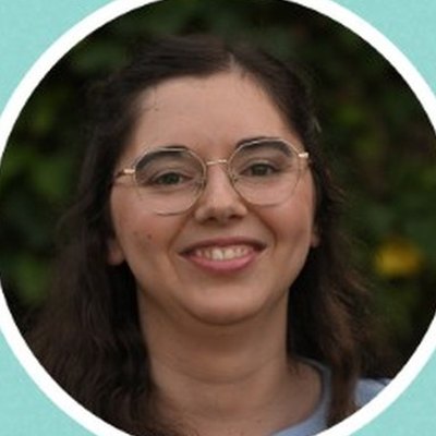 Rita Ferreira, de óculos redondos de armação fina e cabelo escuro, sorri diante de folhagem verde.