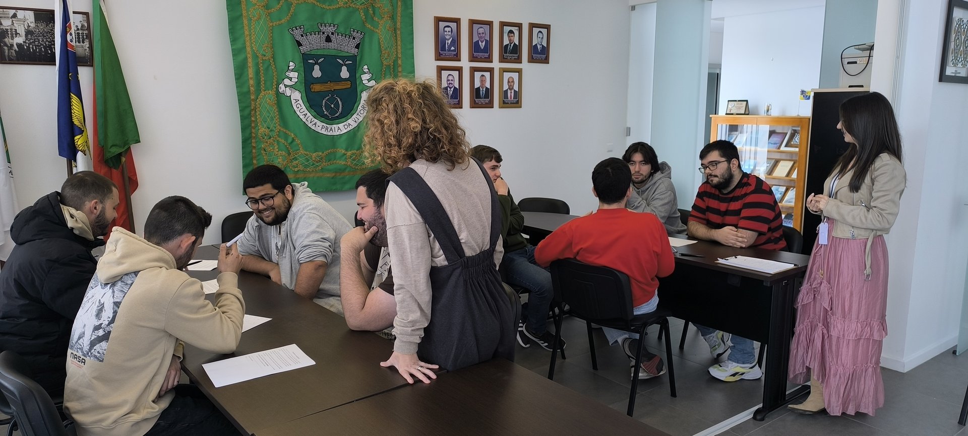 Jovens trabalham em grupo à volta de mesas, com folhas de papel, enquanto uma técnica acompanha de pé.